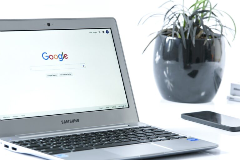 Laptop mit Google-Startseite neben schwarzer Pflanzentopf und Smartphone.
