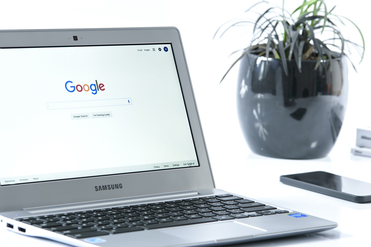 Laptop mit Google-Startseite neben schwarzer Pflanzentopf und Smartphone.