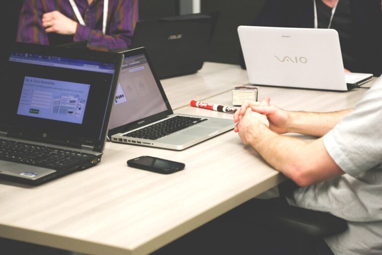 Mehrere Laptops auf Konferenztisch mit zwei Personen im Hintergrund.